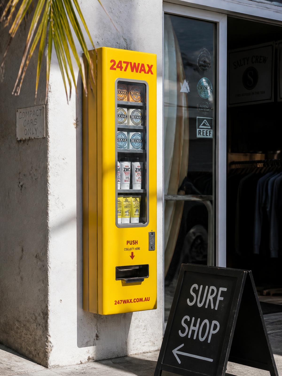 Yellow 247WAX dispenser mounted beside a surf shop entrance, stocked with wax and zinc