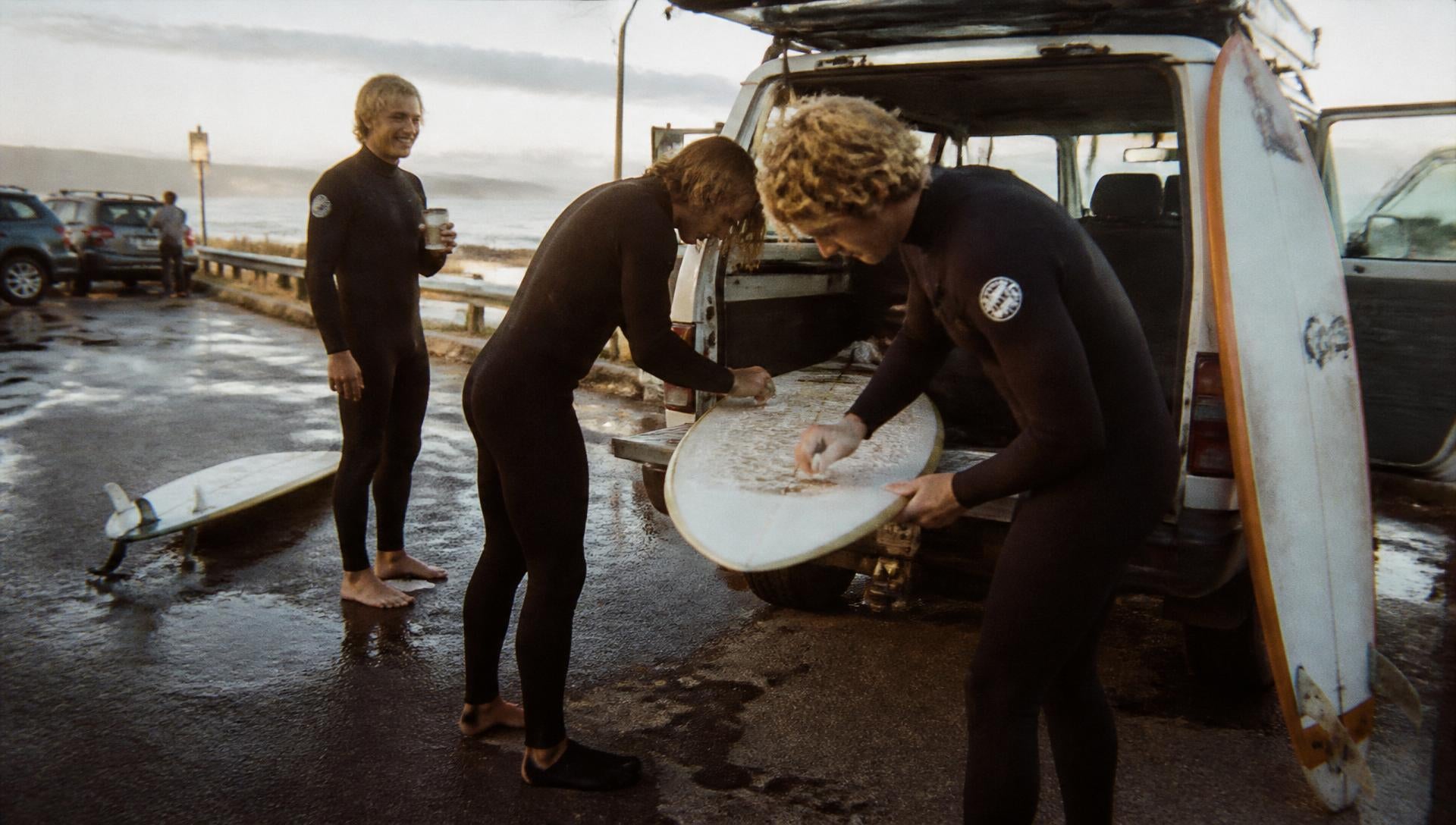 Surfers waxing a board beside their van at a wet, golden-hour carpark before an early surf